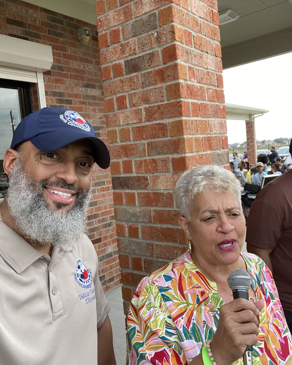 Tahir Charles standing with Councilwoman Dr. Carolyn Evans-Shabazz, who is speaking into a microphone, outside the district Administration Building.