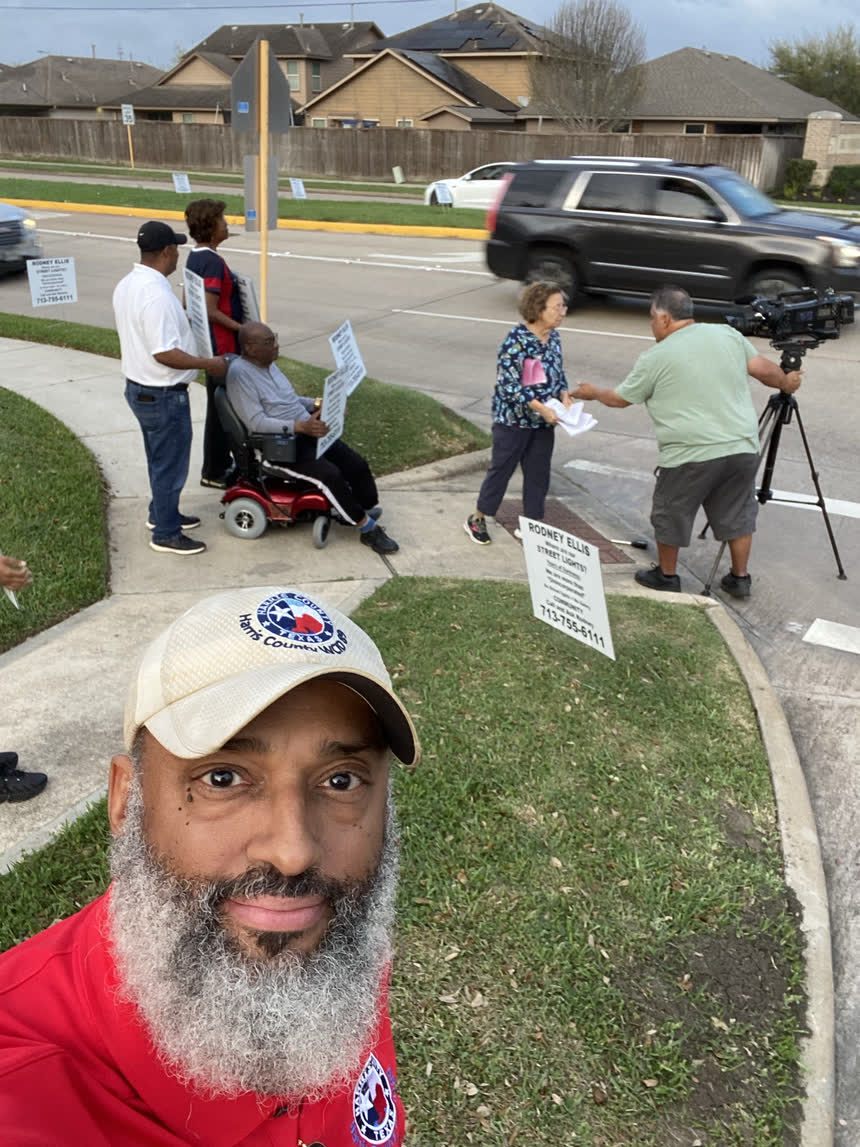 Neighbours holding &ldquo;Where are our street lights?&rdquo; signs on a street corner, one seated in a power wheelchair, while a resident hands papers to a television cameraman.