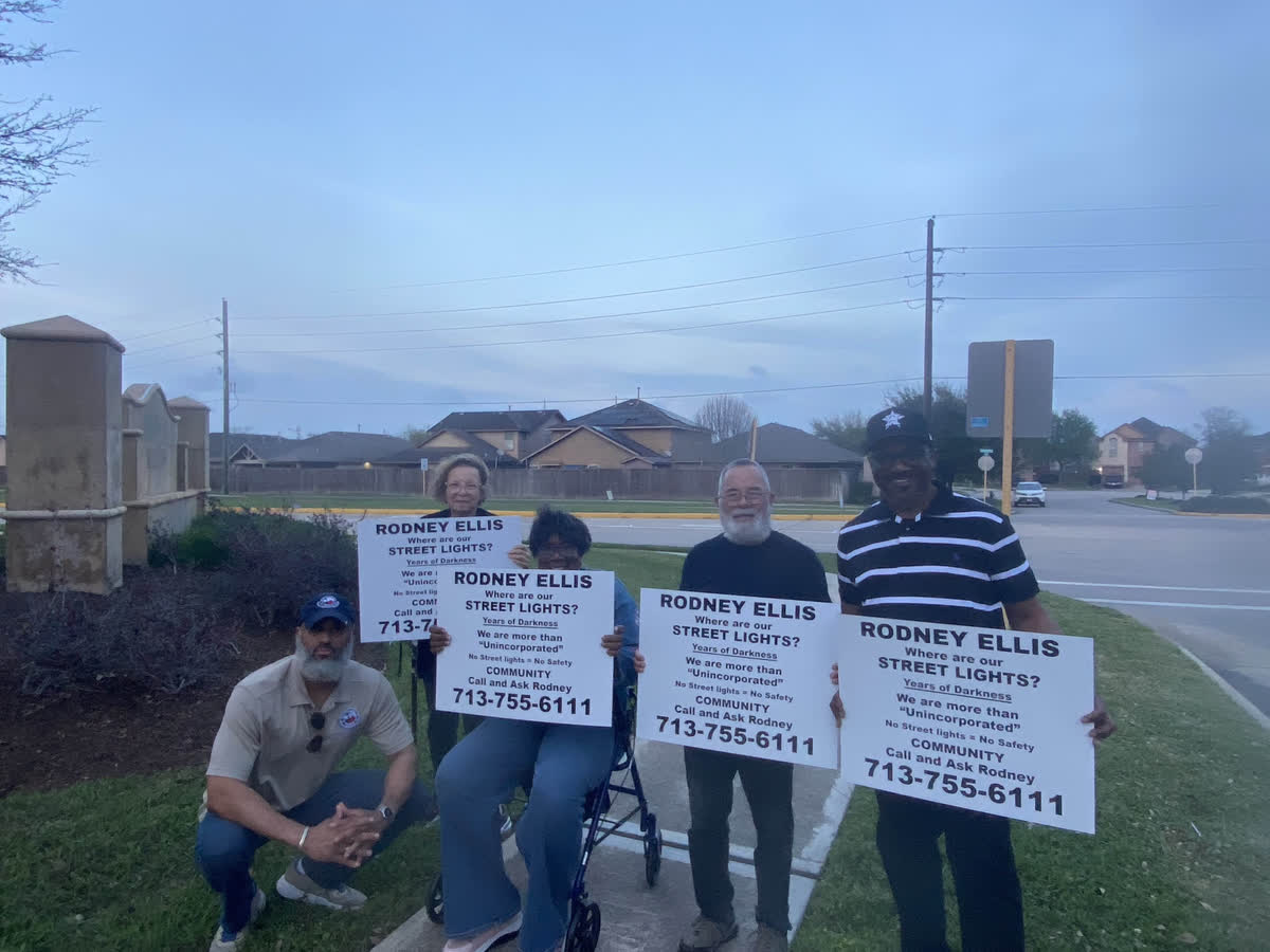 Tahir Charles kneeling beside four neighbours holding &ldquo;Where are our street lights?&rdquo; signs at dusk by the subdivision entrance.
