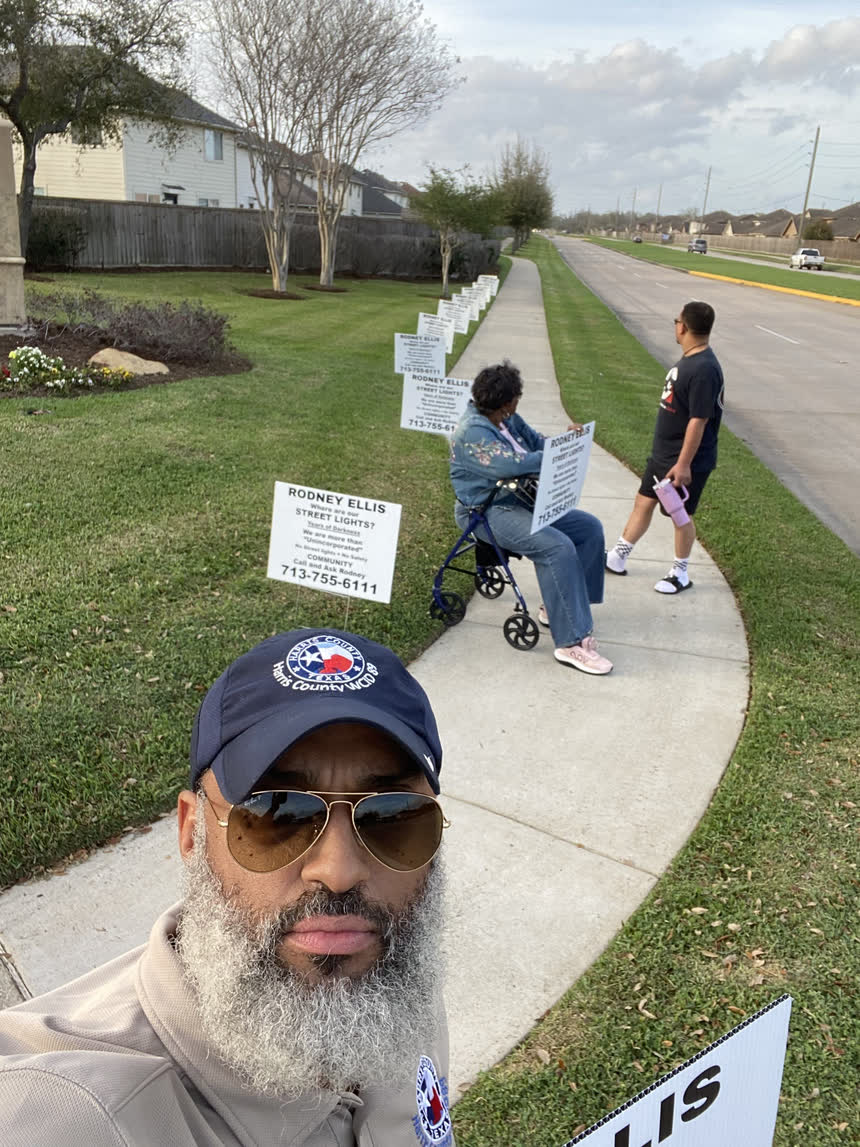 Tahir Charles in a Harris County WCID 89 cap, with a line of &ldquo;Where are our street lights?&rdquo; signs running down the esplanade behind him and neighbours standing along the sidewalk.