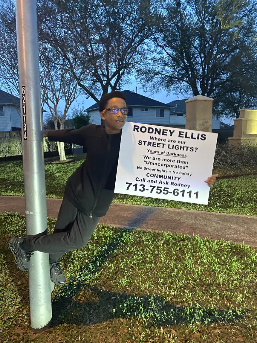 A young resident holding a &ldquo;Where are our street lights?&rdquo; sign while leaning off a pole.