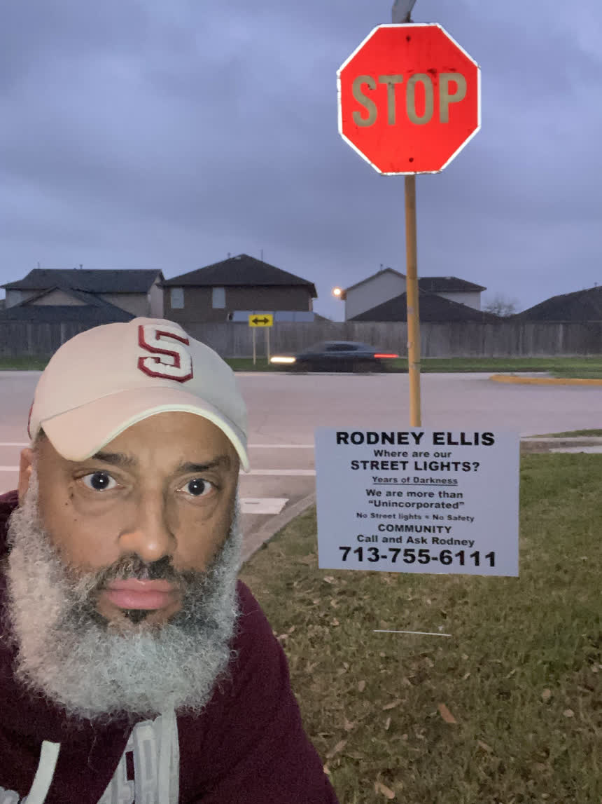 Tahir Charles beside a &ldquo;Where are our street lights?&rdquo; sign planted at a stop sign at dusk, with a car passing behind.